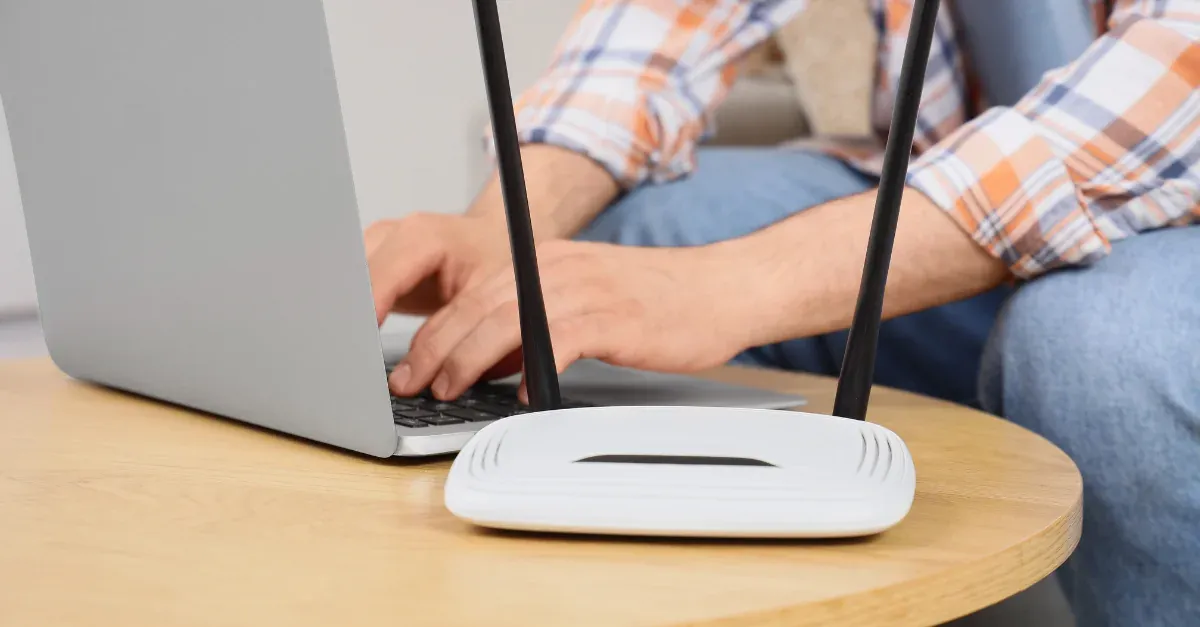 Person typing on a laptop beside a white wireless broadband router on a wooden side table
