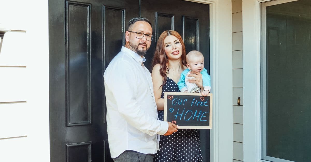 Young family with baby holding a sign 'Our First Home' at doorstep, symbolizing new beginnings.